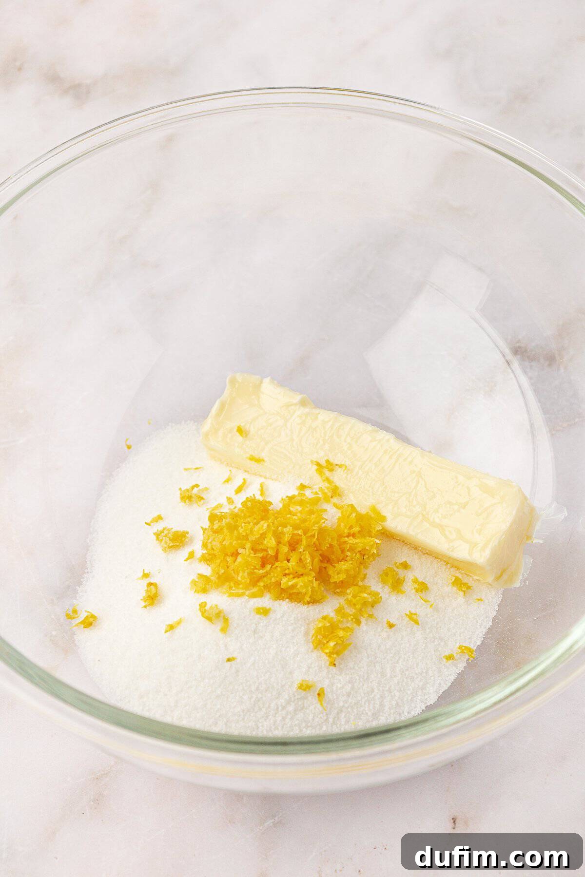 A clear glass bowl containing a stick of butter, granulated sugar, and fresh lemon zest on a marble surface.