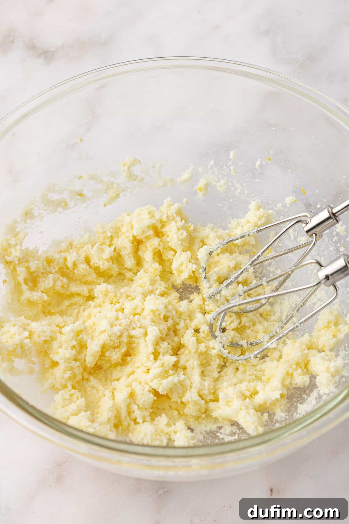A glass bowl containing a creamed mixture of butter and sugar with a hand mixer resting inside, on a light marble surface.