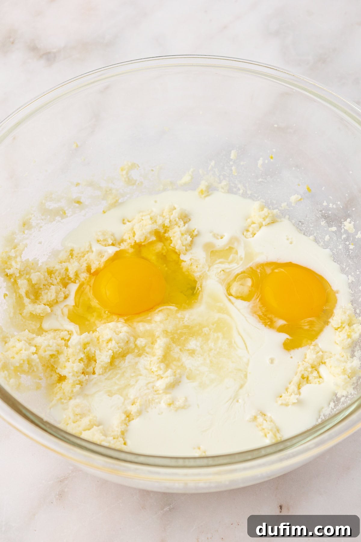 A clear glass bowl contains a mixture of creamed butter and sugar, with two raw eggs and a splash of milk added on top, ready to be mixed together on a light marble surface.