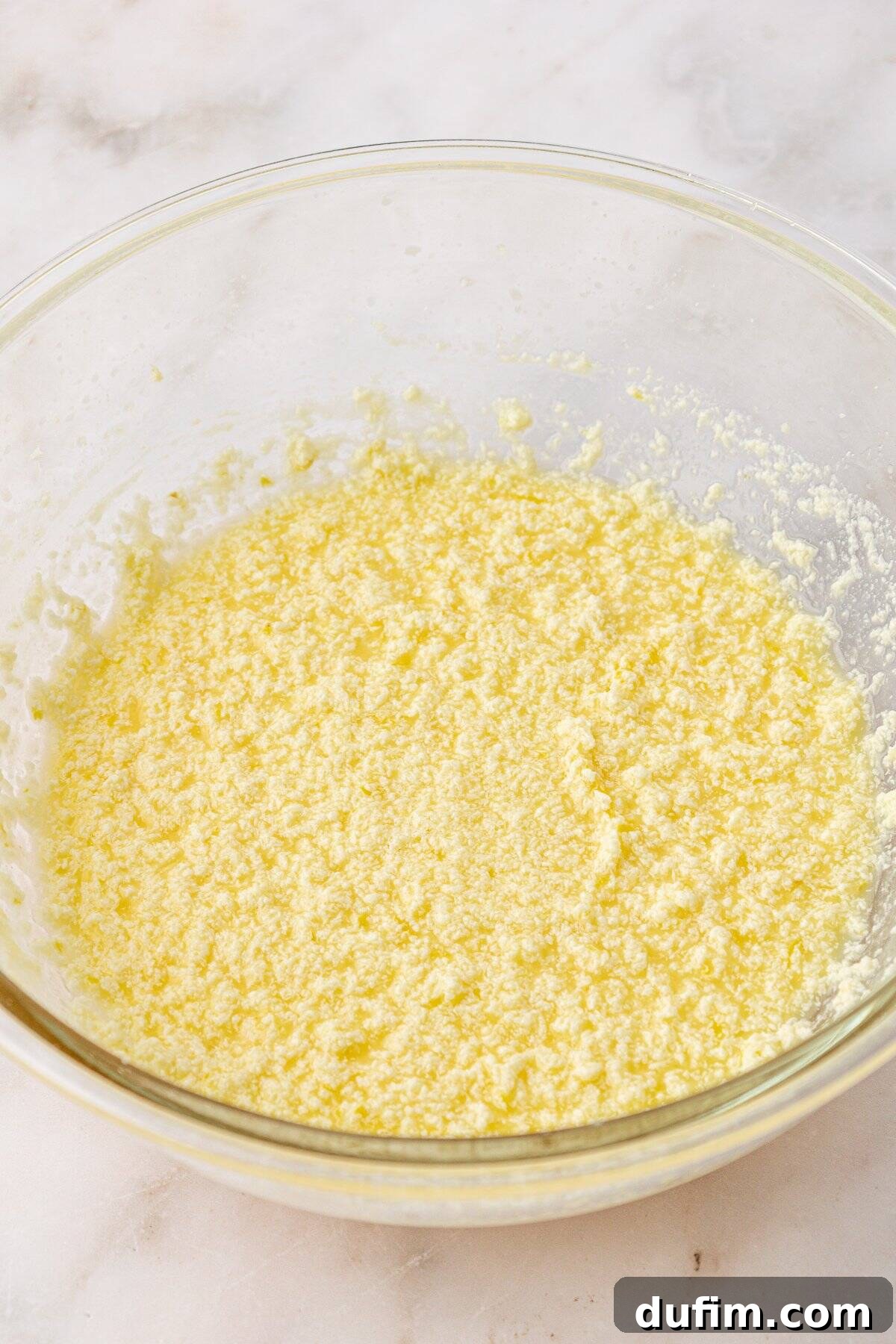 A glass bowl filled with a yellow, grainy mixture sits on a marble surface. The mixture appears to be creamed butter and sugar, often used as a base for baking recipes.