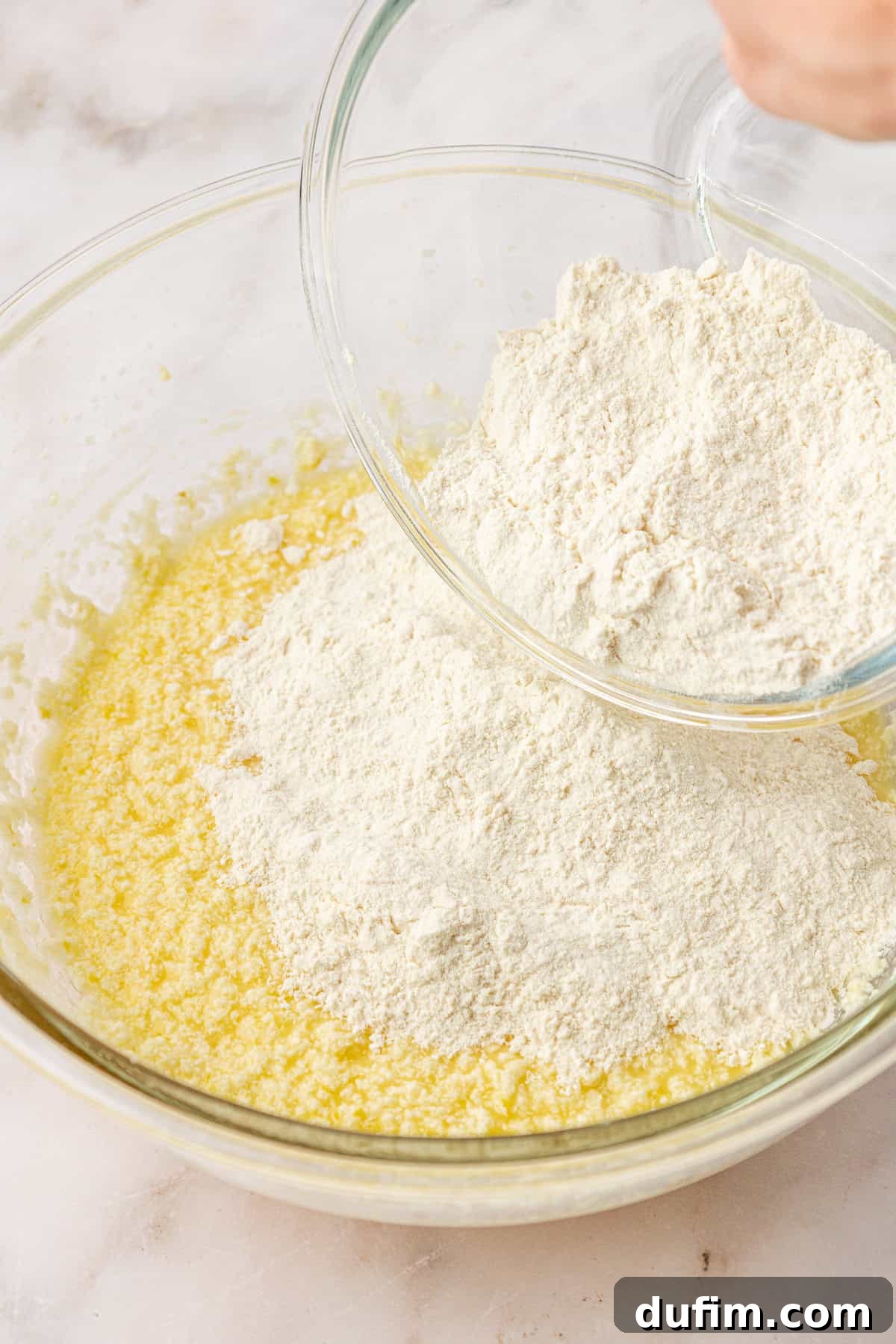 A hand pours flour from a glass bowl into a larger bowl containing a yellow, wet mixture, preparing ingredients for baking on a light-colored surface.