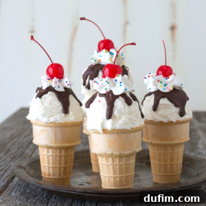 Four ice cream cone cupcakes topped with whipped cream, chocolate sauce, colorful sprinkles, and a cherry, arranged on a round tray on a wooden surface.