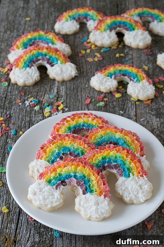 Rainbow Sugar Cookies - use fruity pebbles and shredded coconut to make these delicious rainbow cookies! Five Rainbow Sugar Cookies on a white serving plate with six more rainbow sugar cookie treats in the background on a wooden table.