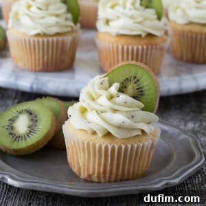 A delicious kiwi cupcake with white frosting and a fresh slice of kiwi sits on a silver plate. Other kiwi cupcakes and pieces of fruit are visible in the background on a marble platter.