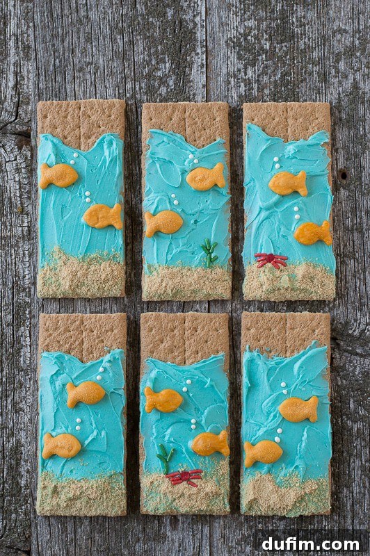 Under the Sea Graham Crackers - easy to make and perfect for an under the sea party! Six vibrant Under the Sea Graham Crackers arranged artfully on a rustic wooden table, adorned with blue buttercream frosting and playful goldfish crackers. They evoke a serene underwater scene.