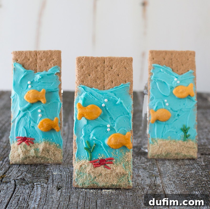 Under the Sea Graham Crackers - easy to make and perfect for an under the sea party! Three delightful Under the Sea Graham Crackers, each beautifully decorated with smooth blue buttercream frosting and charming goldfish crackers, stand upright on a rustic wooden table, ready to be enjoyed.