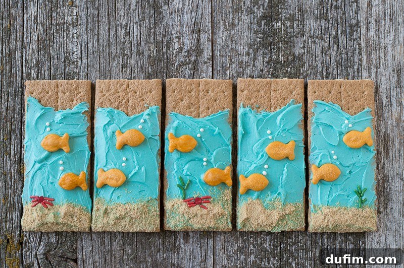 Under the Sea Graham Crackers - easy to make and perfect for an under the sea party! Five beautifully decorated Under the Sea Graham Crackers, adorned with blue buttercream and playful goldfish crackers, are neatly displayed on a wooden table, creating a delightful underwater tableau.
