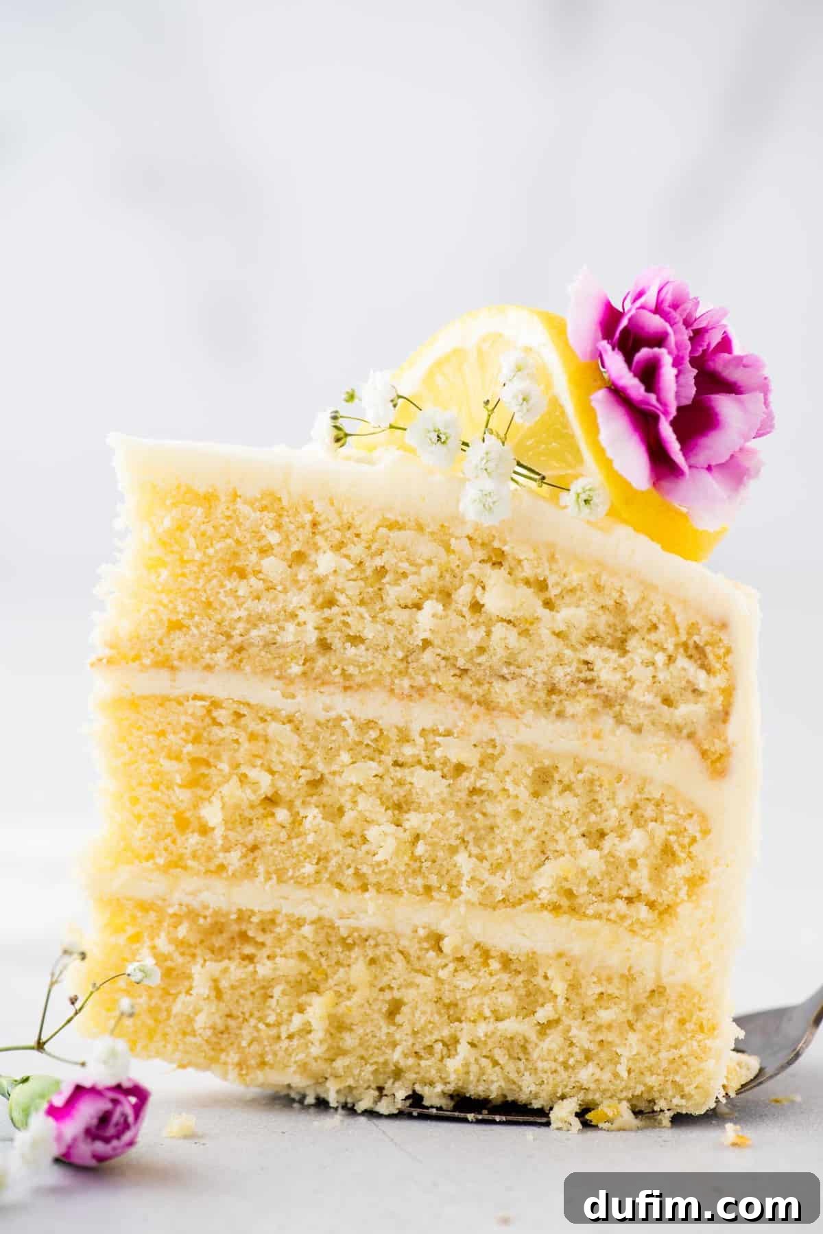 A perfectly plated slice of lemon layer cake on a cake server, elegantly garnished with delicate flowers and fresh lemon slices.