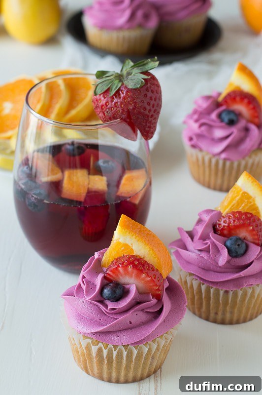 Sangria Cupcakes and a glass of sangria – a match made in heaven Two homemade Sangria Cupcakes with red wine buttercream garnished with blueberries, a slice of orange, and strawberries, positioned next to a glass of sangria. A perfect pairing.