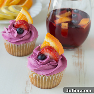 Two sangria cupcakes with purple frosting, each topped with a slice of orange, a slice of strawberry, and a blueberry, sit next to a glass of red fruit punch with visible fruit slices on a white surface.