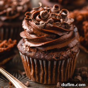 A close-up of rich chocolate cupcakes topped with swirled chocolate frosting and chocolate shavings, with another cupcake blurred in the background.