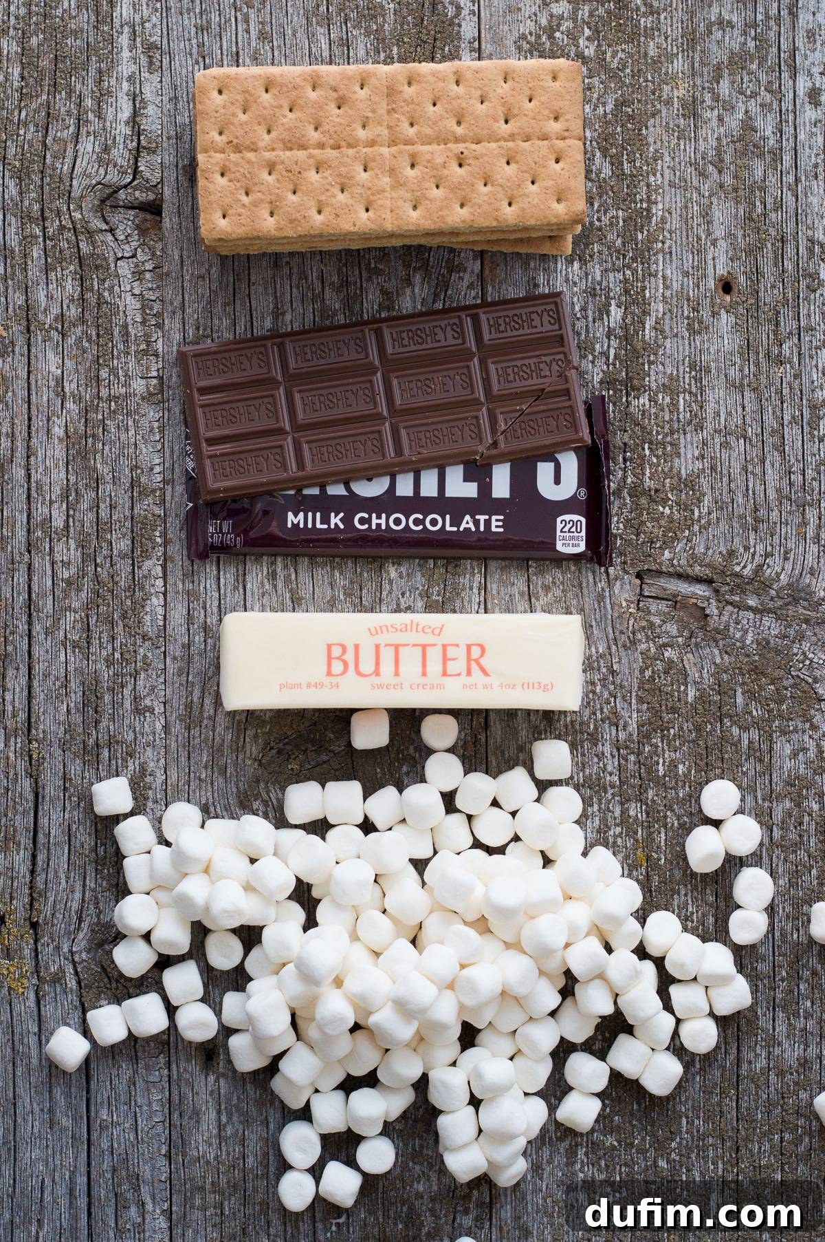 Raw ingredients for S'mores Bars: graham crackers, Hershey's chocolate bars, a stick of butter, and a bag of mini marshmallows, artfully arranged on a rustic gray wood background.