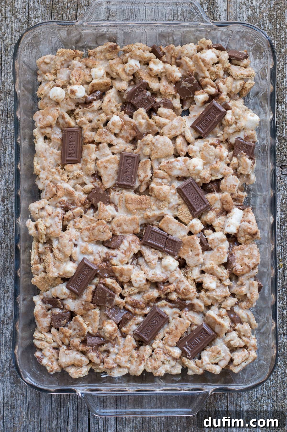 S'mores bars setting in a glass 9x13 inch pan, topped with additional Hershey's chocolate bar pieces, ready for cooling and cutting.