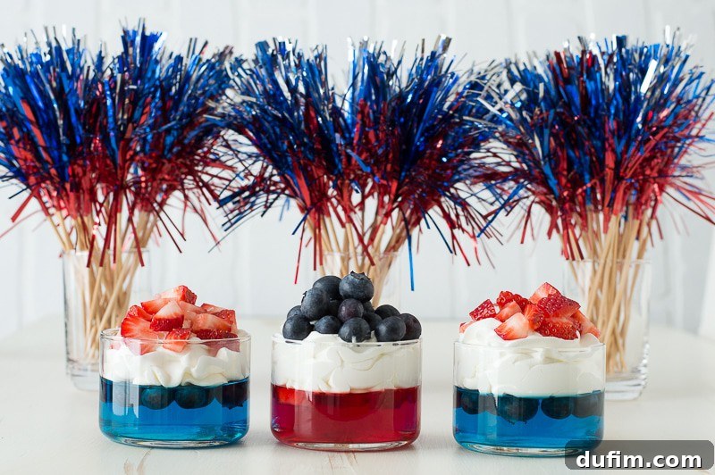 A vibrant display of red and blue jello cups, each generously topped with a cloud of whipped cream and garnished with a mix of fresh, juicy strawberries and plump blueberries. The desserts are arranged artfully, inviting guests to indulge in a festive treat.