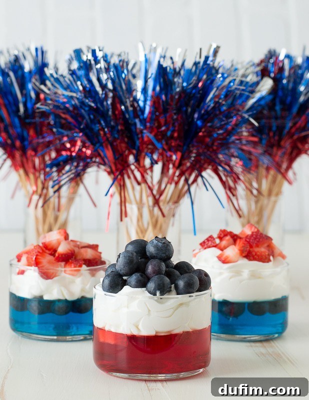 Four individual glass cups displaying beautiful red and blue jello layers, each generously topped with a swirl of whipped cream and garnished with fresh strawberries and blueberries. The desserts are arranged on a festive, subtly textured surface, creating an inviting and celebratory atmosphere, perfect for any holiday gathering.