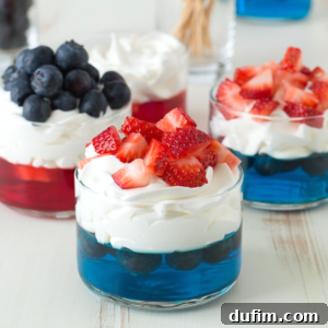Three glass cups with festive 4th of July Jello cups: blue gelatin layered with blueberries and whipped cream, one topped with extra blueberries, while the others have strawberries on whipped cream over red or blue gelatin.
