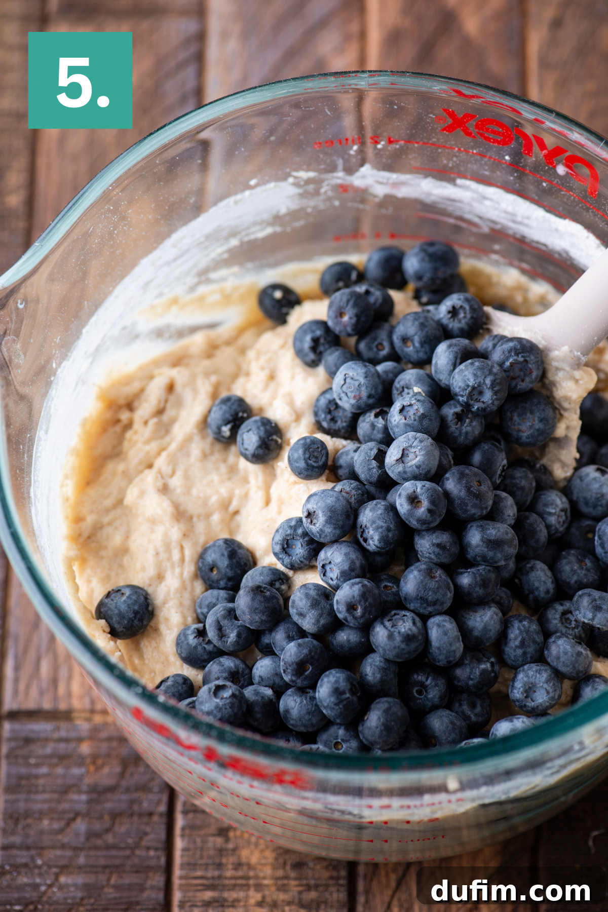 Wholesome Blueberry Muffins 11 A glass mixing bowl filled with muffin batter and fresh blueberries, being stirred with a white spatula, sits on a wooden surface. A green box with the number 5 appears in the top left corner.
