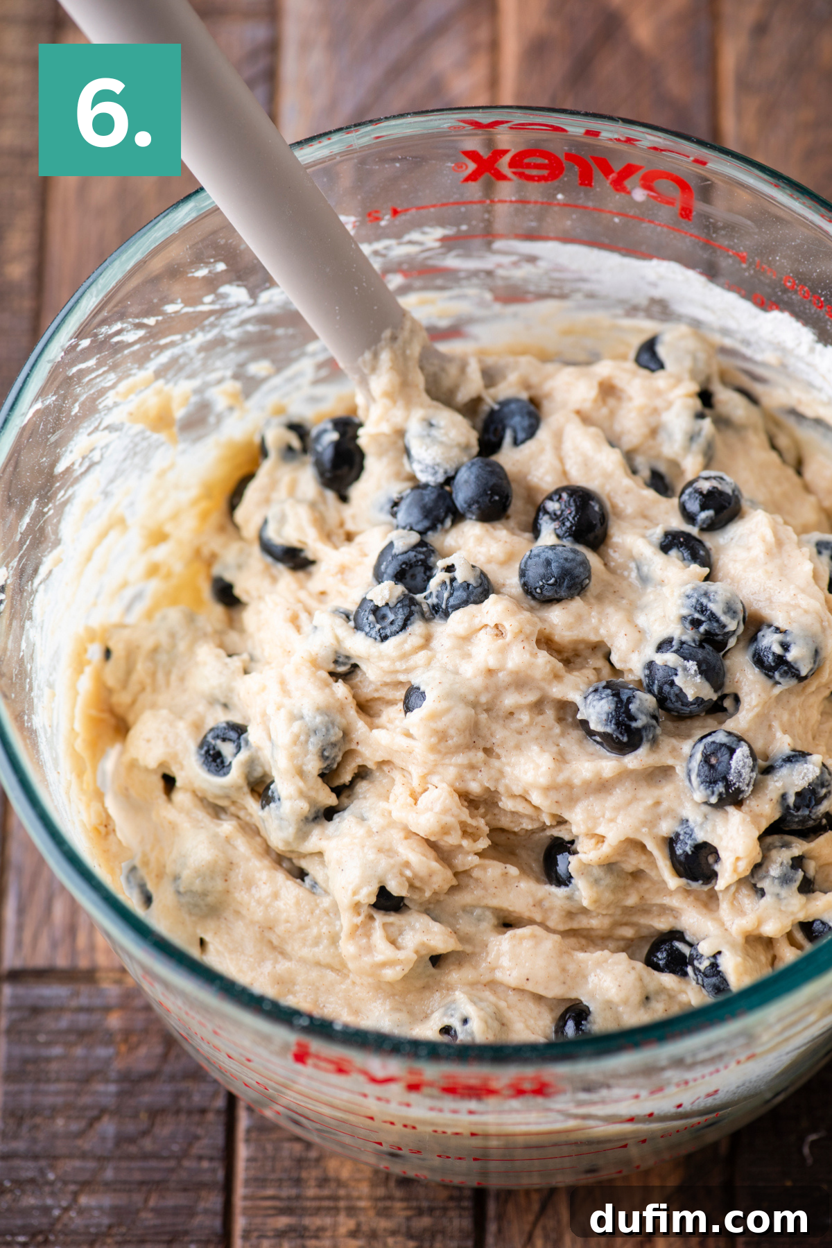 Wholesome Blueberry Muffins 12 A glass mixing bowl filled with thick blueberry muffin batter and a spatula. The bowl sits on a wooden surface, and a green square with the number 6 is in the top left corner.