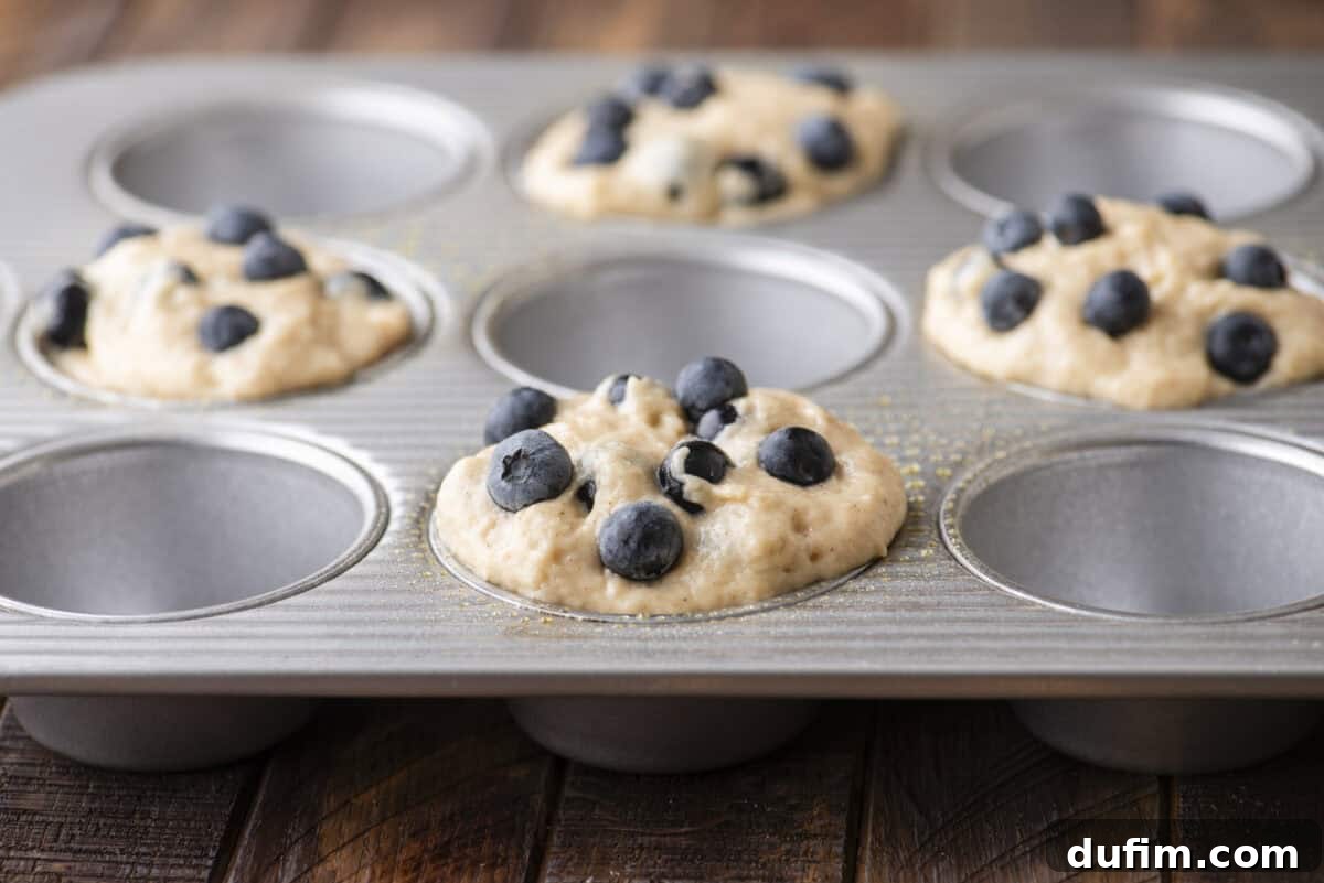 Wholesome Blueberry Muffins 13 A metal muffin tin with several cups filled with blueberry muffin batter, some topped with fresh blueberries, resting on a wooden surface. A few cups are empty.
