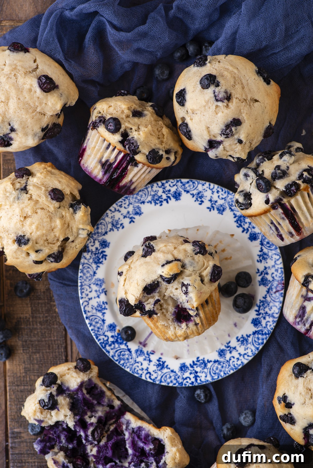 Wholesome Blueberry Muffins 15 Overhead view of several blueberry muffins, some on a blue-patterned plate and others on a wooden surface and blue cloth, with loose blueberries scattered around. One muffin is cut open, showing the blueberry-filled center.