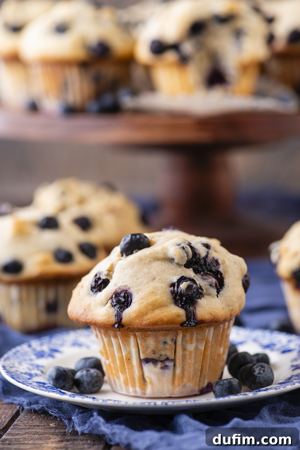 Wholesome Blueberry Muffins 3 A freshly baked healthy blueberry muffin sits on a blue and white plate with whole blueberries, with more muffins on a cake stand in the background. The muffin is golden brown and studded with juicy blueberries.