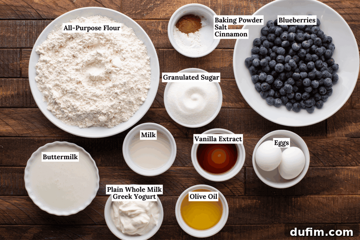 Wholesome Blueberry Muffins 6 Various baking ingredients in bowls on a wooden surface, including all-purpose flour, granulated sugar, buttermilk, milk, Greek yogurt, olive oil, vanilla extract, eggs, blueberries, and a mix of baking powder, salt, and cinnamon.