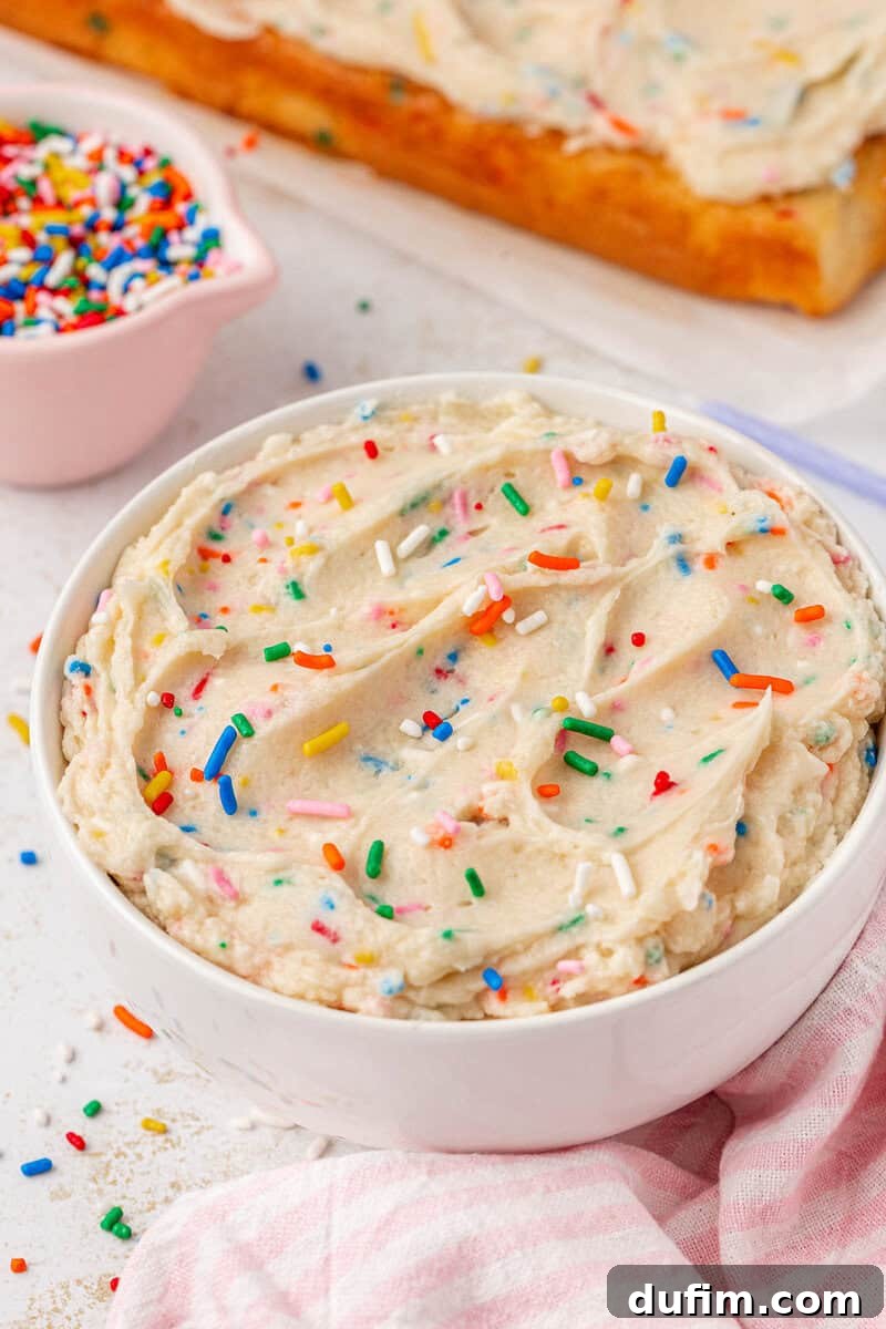 Effortlessly Light and Fluffy Funfetti Frosting 2 A white bowl filled with creamy funfetti frosting topped with colorful rainbow sprinkles sits on a white surface. A pink-striped cloth and a small bowl of extra sprinkles are next to it. A cake with matching frosting is in the background.