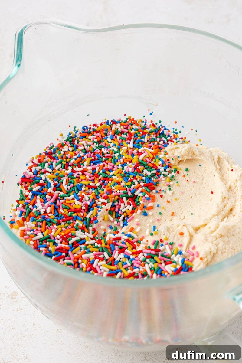 Effortlessly Light and Fluffy Funfetti Frosting 11 A glass mixing bowl with cookie dough and colorful rainbow sprinkles sitting on top, ready to be mixed together.