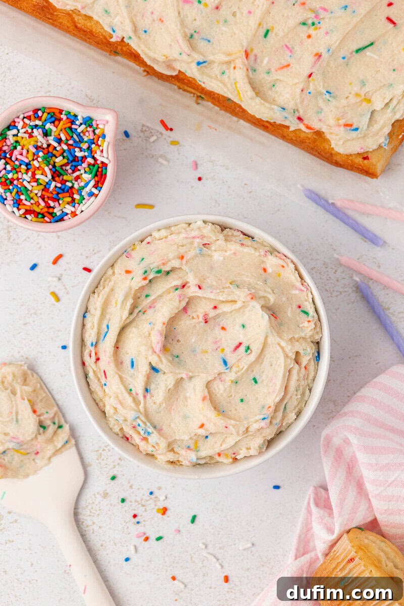 Effortlessly Light and Fluffy Funfetti Frosting 13 A bowl of funfetti frosting with colorful sprinkles sits on a white surface, surrounded by a spatula, a small bowl of extra sprinkles, pink and purple candles, and a partially frosted cake.