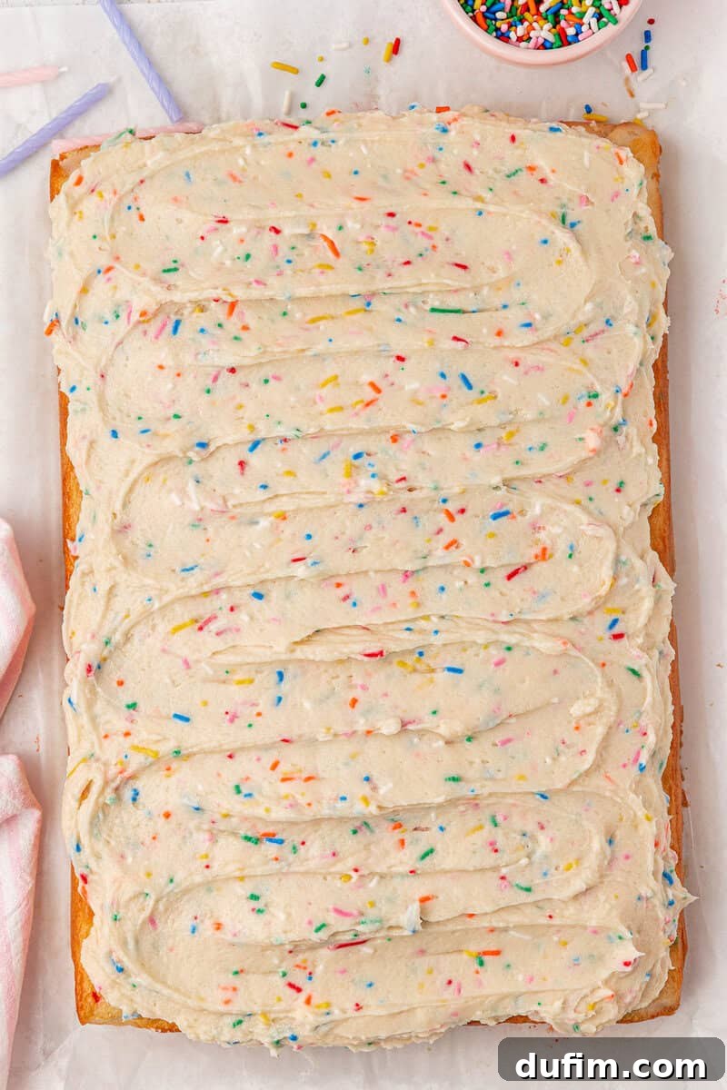 Effortlessly Light and Fluffy Funfetti Frosting 3 A rectangular sheet cake topped with swirled vanilla frosting and colorful rainbow sprinkles on parchment paper, with a bowl of extra sprinkles nearby.