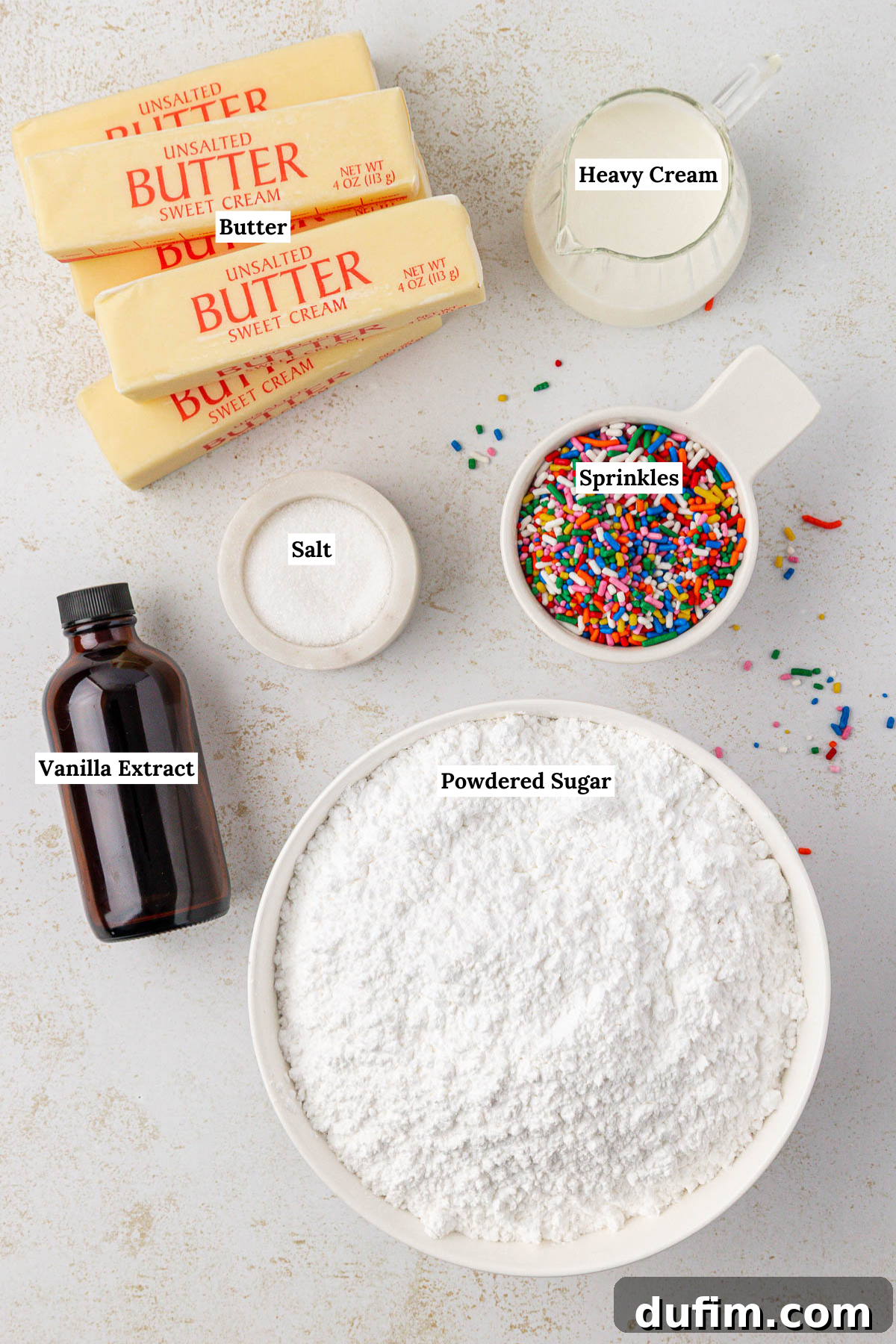 Effortlessly Light and Fluffy Funfetti Frosting 4 Top-down view of baking ingredients: four sticks of butter, a small pitcher of heavy cream, a bowl of sprinkles, a bottle of vanilla extract, a bowl of salt, and a large bowl of powdered sugar, all labeled.