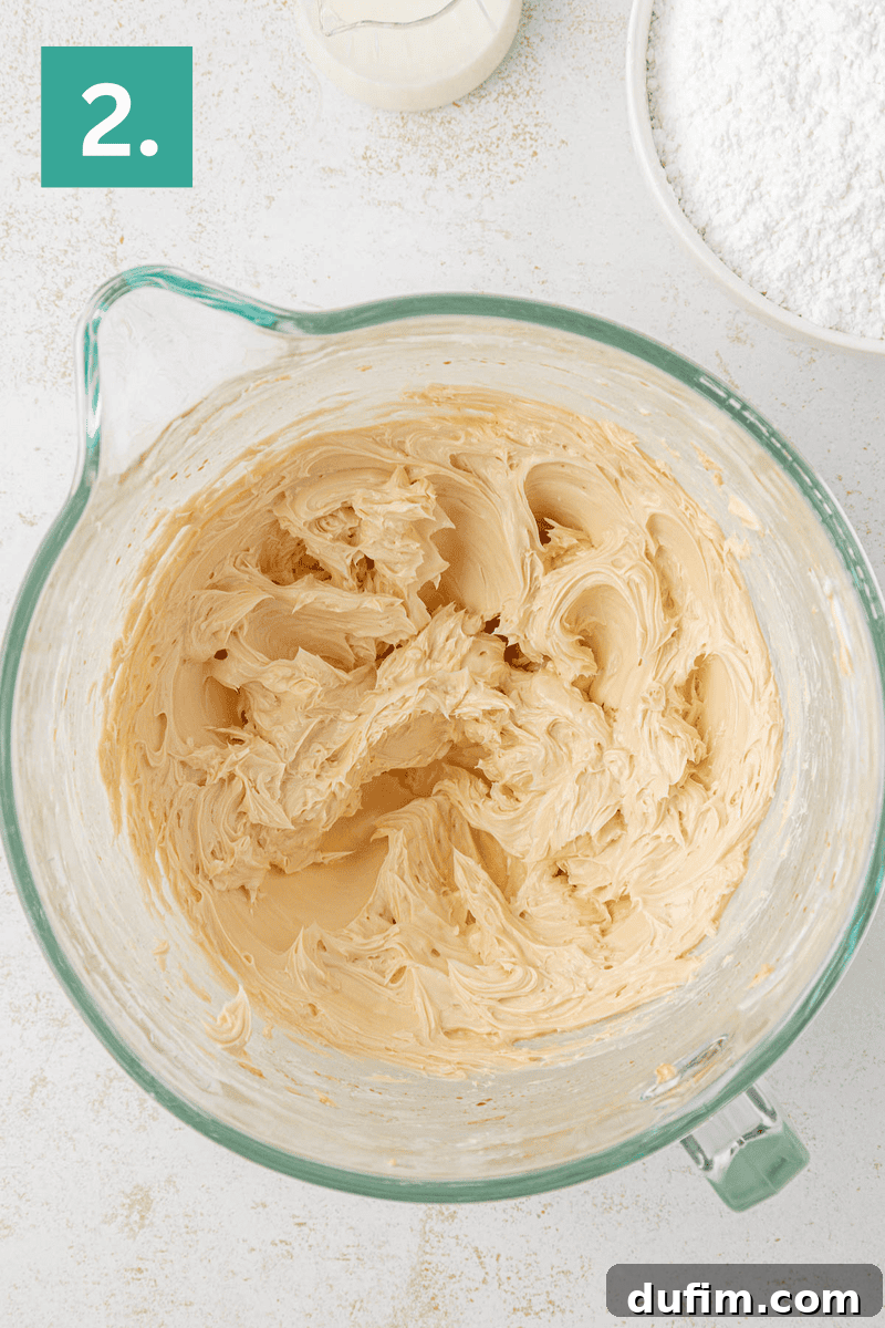 Effortlessly Light and Fluffy Funfetti Frosting 6 A glass mixing bowl filled with creamy, light brown butter mixture. A bowl of powdered sugar and a small bottle of liquid are nearby. A green square in the corner displays the number 2.