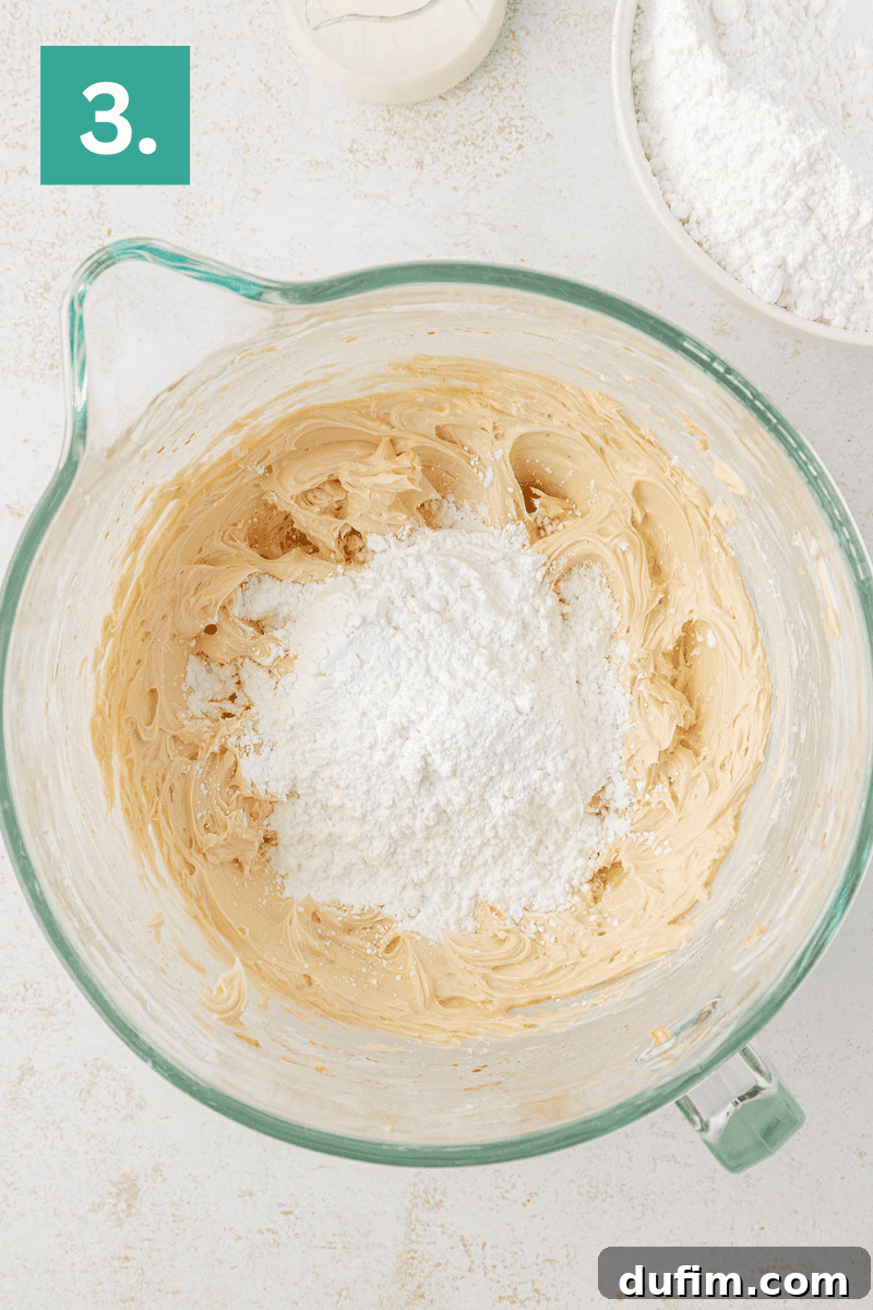 Effortlessly Light and Fluffy Funfetti Frosting 7 A glass mixing bowl with creamy batter and a heap of powdered sugar in the center. Part of a white bowl with flour and a bottle of milk are nearby. A green box with the number 3 appears in the upper left corner.
