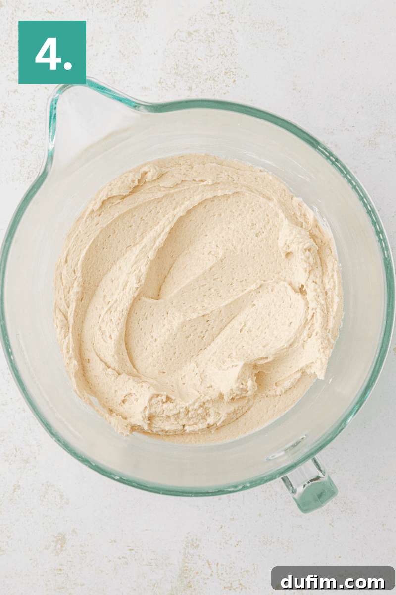 Effortlessly Light and Fluffy Funfetti Frosting 8 A glass mixing bowl filled with creamy, light brown batter sits on a white surface. A green box with the number 4 is in the top left corner.