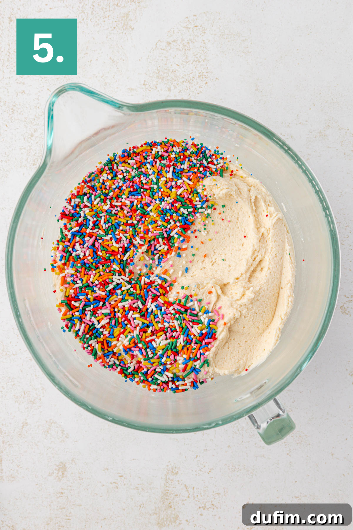 Effortlessly Light and Fluffy Funfetti Frosting 9 A glass mixing bowl filled with creamy cake batter and colorful rainbow sprinkles on top, set on a light surface. A green square with a white number 5 is in the top left corner.
