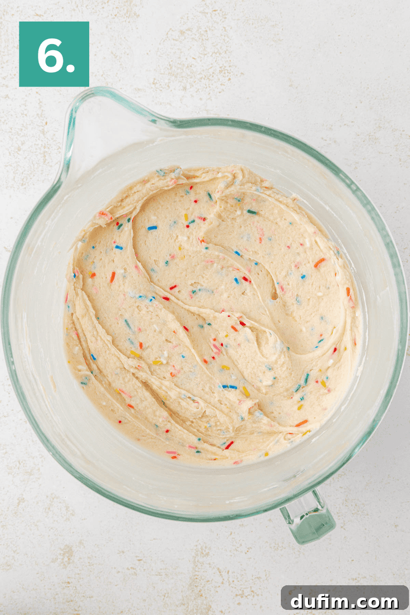 Effortlessly Light and Fluffy Funfetti Frosting 10 A glass mixing bowl filled with creamy cake batter mixed with colorful sprinkles. The bowl is placed on a light surface, and a green box with the number 6 is in the top left corner.