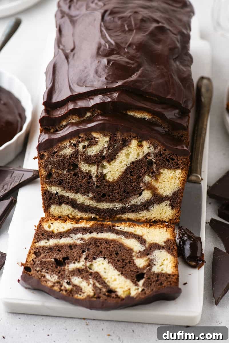 A beautifully sliced marble loaf cake with distinct chocolate and vanilla swirls, generously topped with a glossy chocolate icing, resting on a pristine white cutting board alongside a knife and decorative chocolate pieces.