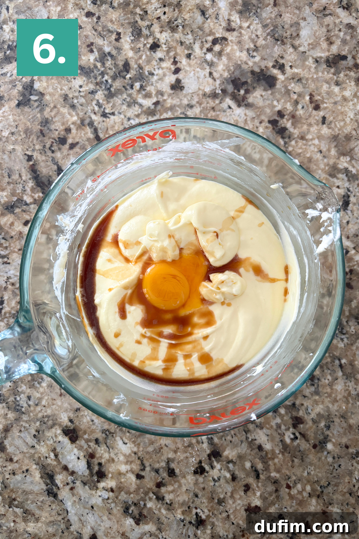 A creamy, pale yellow batter in a glass mixing bowl, featuring a whole egg, a dollop of butter, and vanilla extract on top, awaiting final blending.