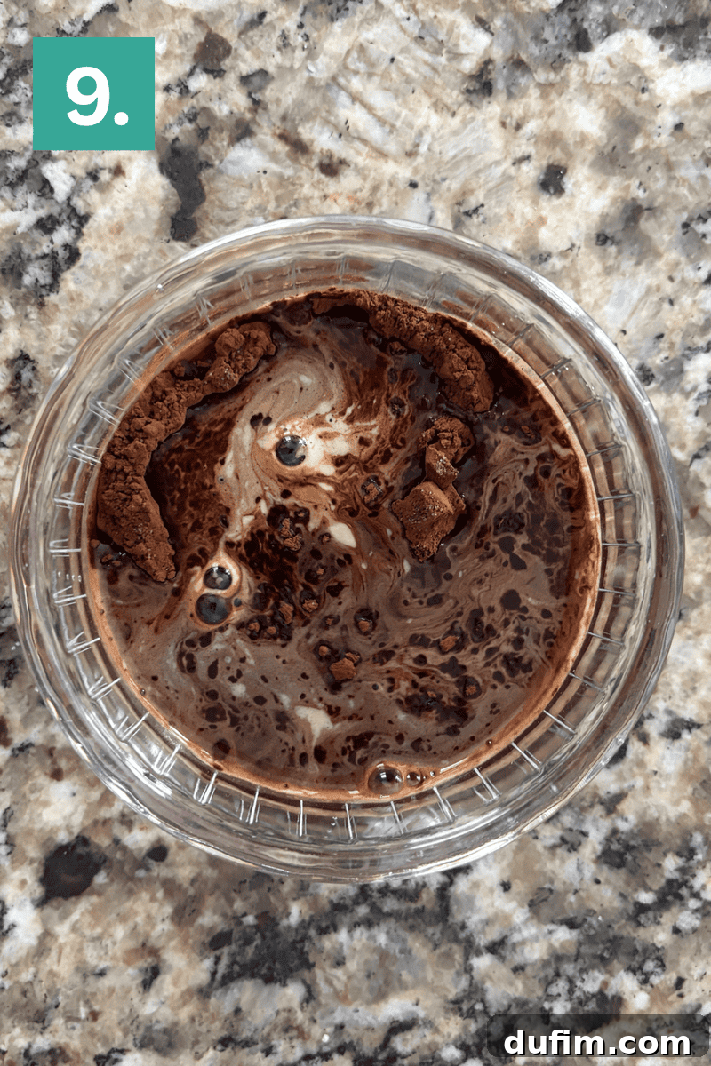 A clear glass bowl containing a rich, dark cocoa powder swirled with a hot liquid, forming a thick, pudding-like mixture, ready to be added to batter.