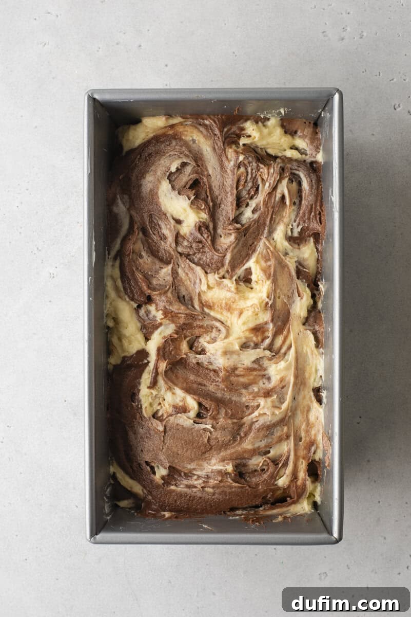 A rectangular metal baking pan containing raw chocolate and vanilla marbled cake batter, skillfully swirled into an appealing pattern, awaiting its transformation in the oven.