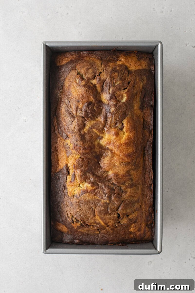 A freshly baked loaf of banana bread with a rich, golden-brown and slightly cracked top, resting in a rectangular metal baking pan on a light gray surface, cooling down.