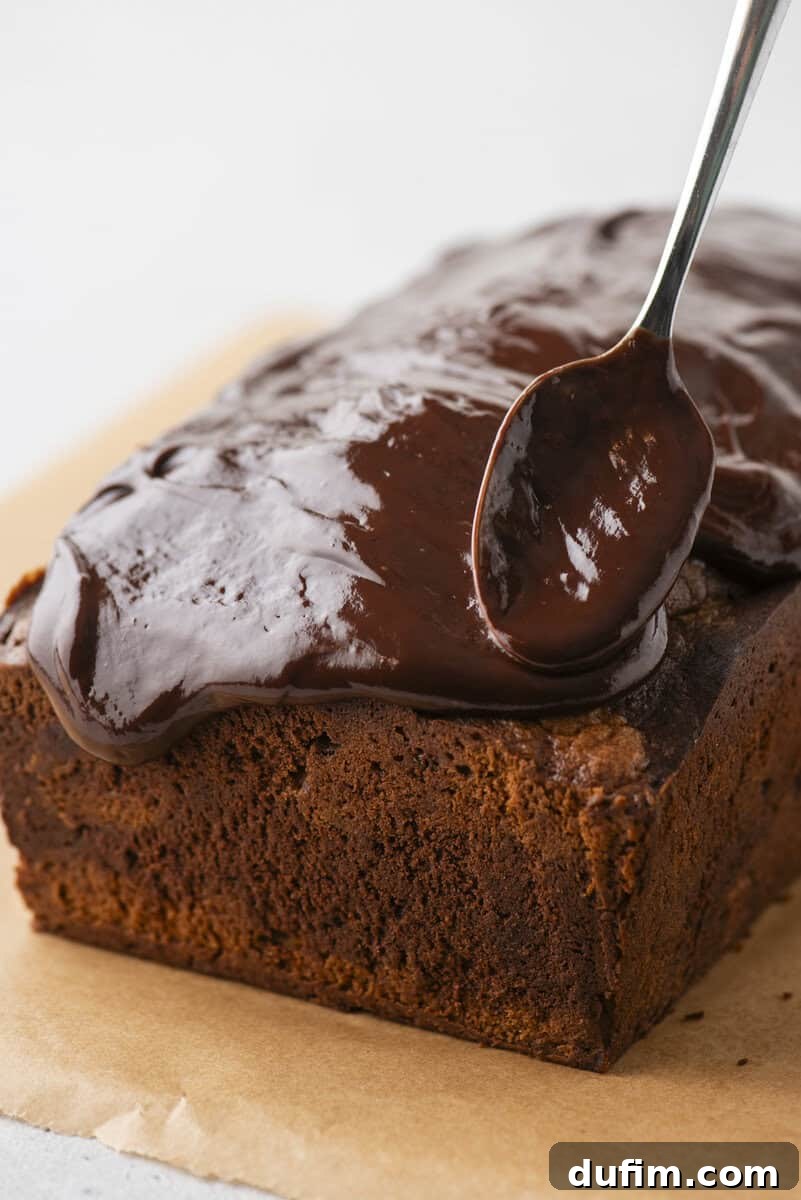 A close-up view of a baked chocolate cake loaf on parchment paper, being artfully drizzled and spread with a spoonful of glossy chocolate icing.