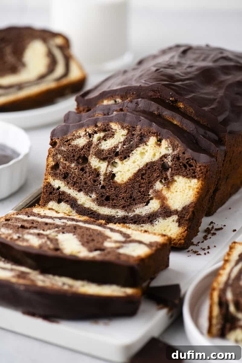 A perfectly sliced marble loaf cake, featuring intricate swirls of chocolate and vanilla, is crowned with a smooth, glistening layer of chocolate glaze. It rests invitingly on a white cutting board, beside a refreshing glass of milk.