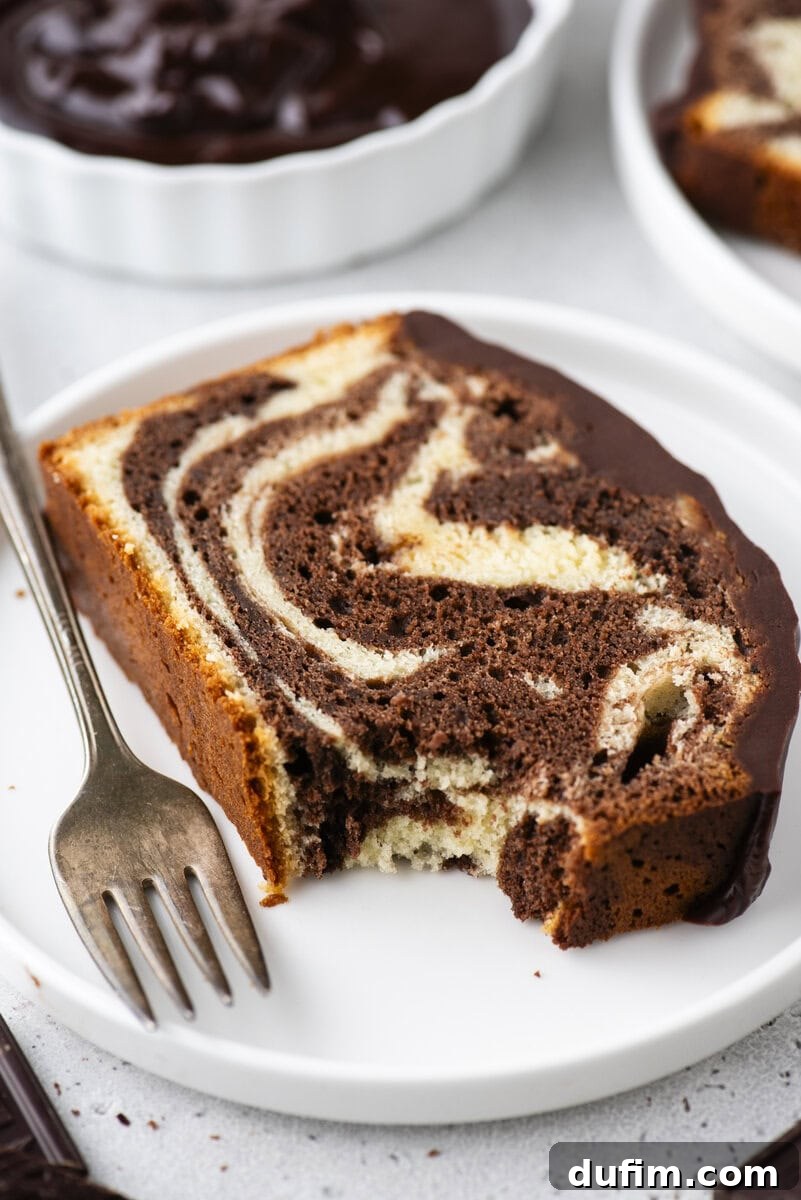 A delectable slice of marble cake, glistening with a smooth chocolate glaze, is elegantly presented on a white plate beside a fork. A perfect bite has been taken, revealing the intricate swirls of chocolate and vanilla within its tender crumb.