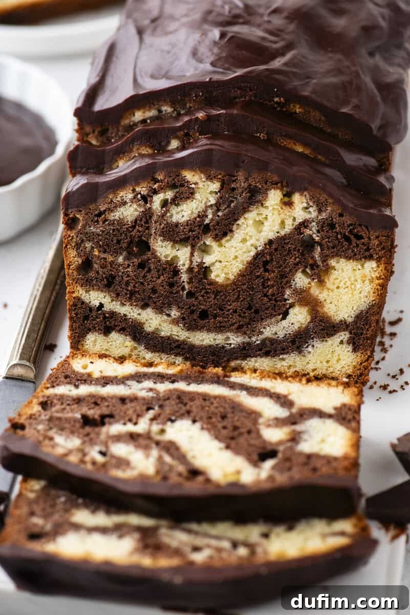 A slice of elegant marble pound cake, showcasing its beautiful swirls of vanilla and chocolate, is topped with a generous, glossy layer of chocolate glaze, all presented on a pristine white surface with a serving knife close by.