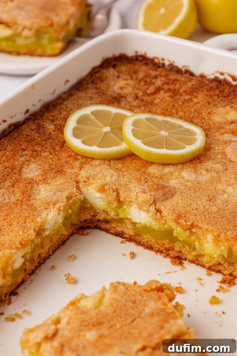 A perfectly baked lemon dessert with a shimmering golden crust sits in a white rectangular baking dish. Two fresh lemon slices garnish the top, and a neat square slice has been removed, revealing layers of gooey lemon filling and creamy pockets inside.