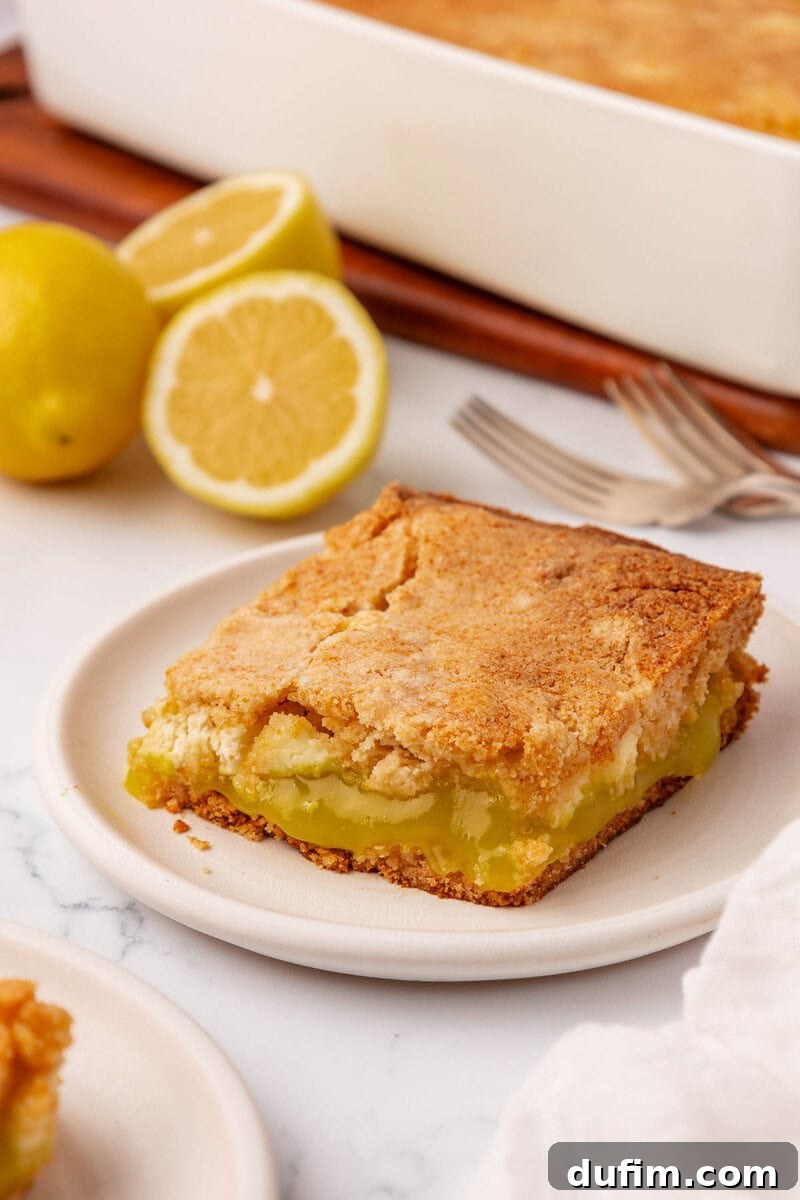 A decadent slice of lemon dessert bar, featuring a perfectly golden, crumbly top, is elegantly presented on a white plate. In the background, a pristine white baking dish holds the remaining dessert, accompanied by two freshly cut lemons and two forks, all resting on a luxurious white marble surface.