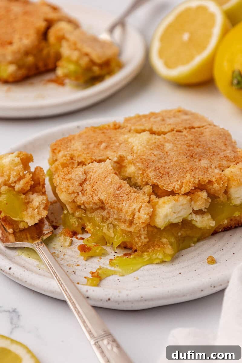 A close-up photograph captures a luscious slice of lemon dessert, distinguished by its crumbly golden crust and sumptuously gooey lemon filling, presented elegantly on a ceramic plate. A silver fork rests nearby, inviting a taste, with fresh lemon halves artfully blurred in the background, hinting at the cake's vibrant citrus notes.