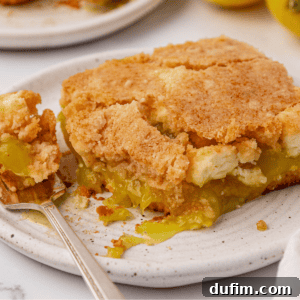 A close-up of a slice of lemon pie with a crumbly golden crust and gooey lemon filling on a white plate; a fork rests beside the pie holding a small bite.