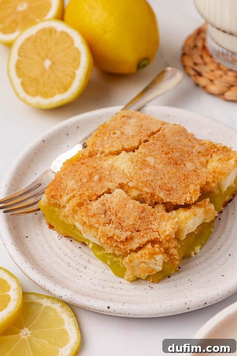 A perfectly portioned square slice of a lemon bar, featuring a rich, crumbly golden crust, rests gracefully on a crisp white plate beside a gleaming fork. In the soft-focus background, an abundance of fresh lemon halves and whole lemons adds a vibrant, citrusy touch to the scene.